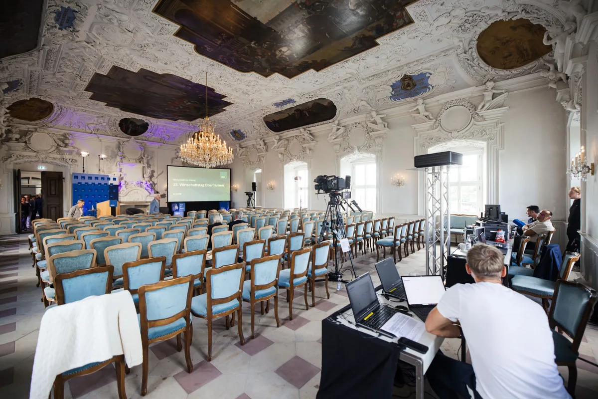 Noch leerer Saal mit blau gepolsterten Stühlen unter Stuckdecke und Kronleuchter, vorn ein Techniker an Laptops