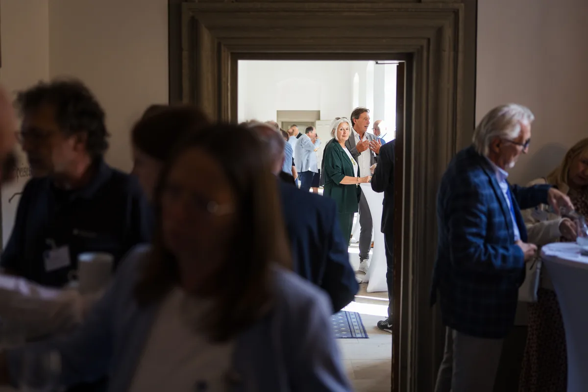 Blick durch einen steinernen Türrahmen auf plaudernde Gäste im Nebenraum, im Vordergrund unscharfe Köpfe und Schultern