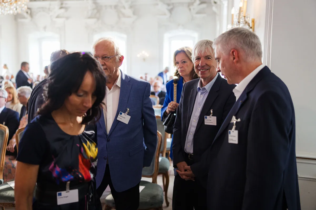 Gäste mit Namensschildern stehen im Saal und blicken nach unten, hinter ihnen besetzte Stuhlreihen vor hohen Fenstern