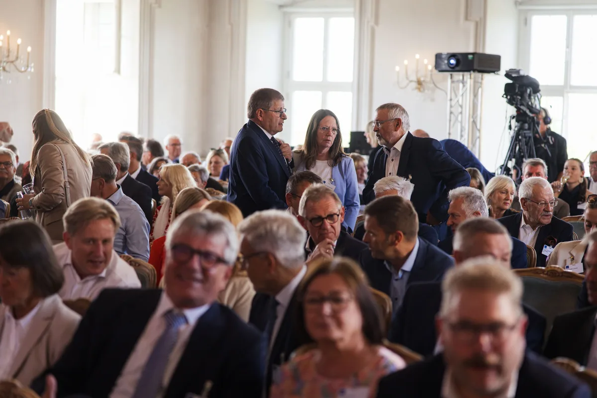 Zuhörer in dichten Stuhlreihen, in der Mitte stehen ein Mann im dunklen Anzug und eine Frau im Gespräch, hinten eine Kamera