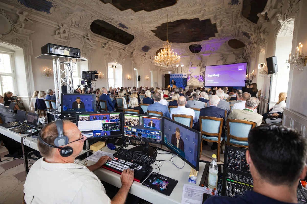 Techniker mit Kopfhörer am Regiepult vor mehreren Monitoren, dahinter das Publikum und das Podium am Ende des Saals