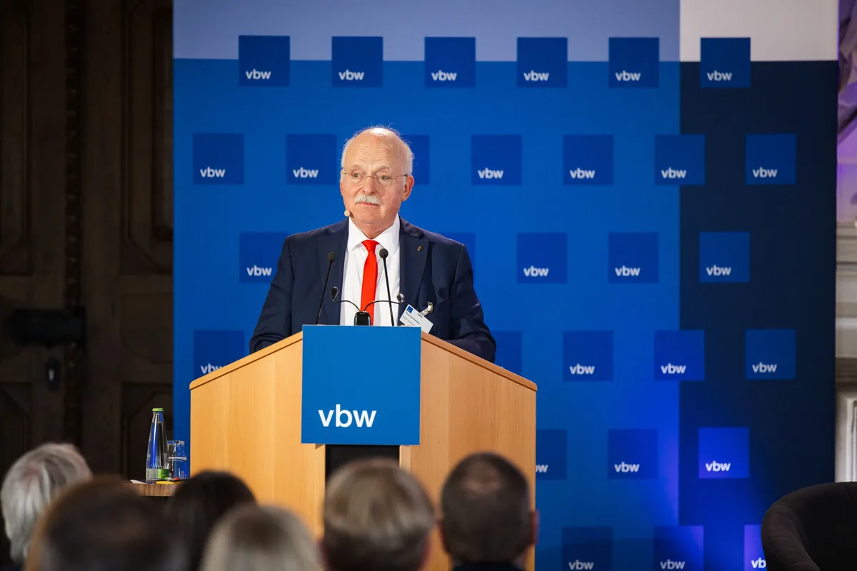 Älterer Redner mit Schnauzbart, Brille und roter Krawatte spricht am Pult vor der blauen Wand, davor unscharf Zuhörer