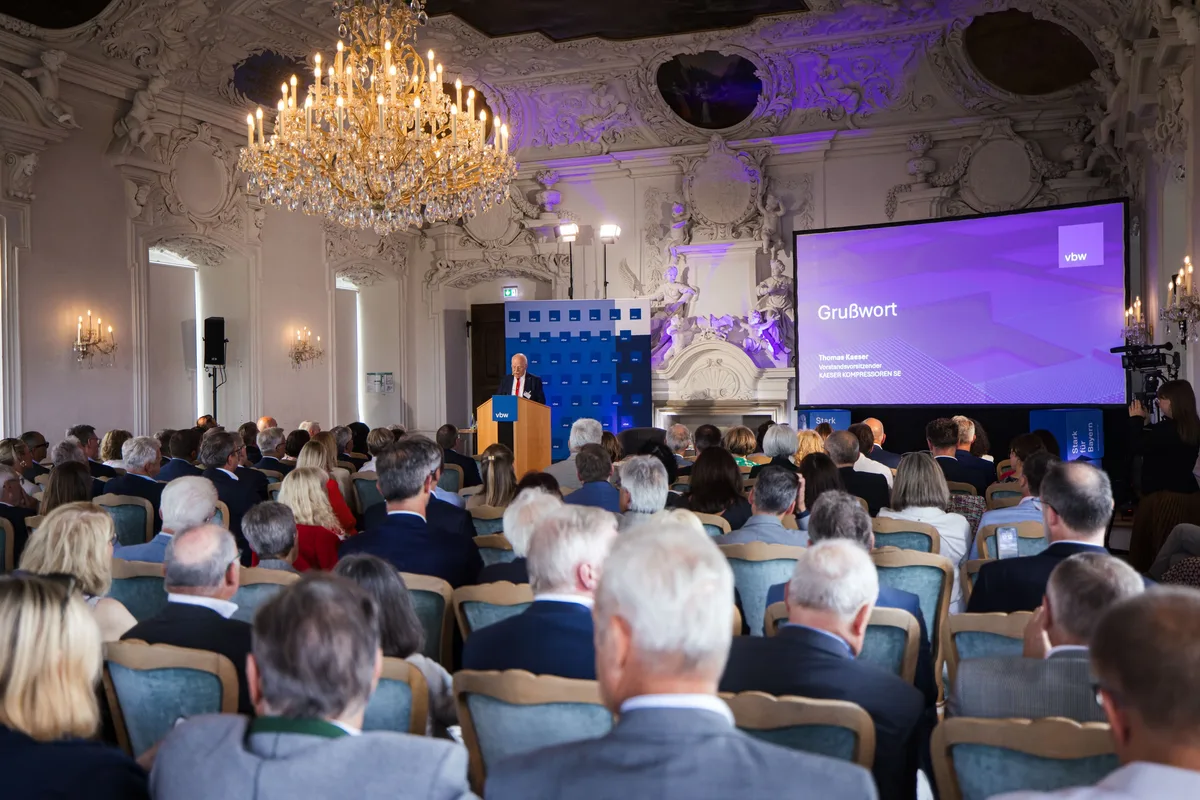 Saal von hinten mit Kronleuchter und Stuckdecke, vorn spricht ein Redner am Pult, rechts die Leinwand Grußwort