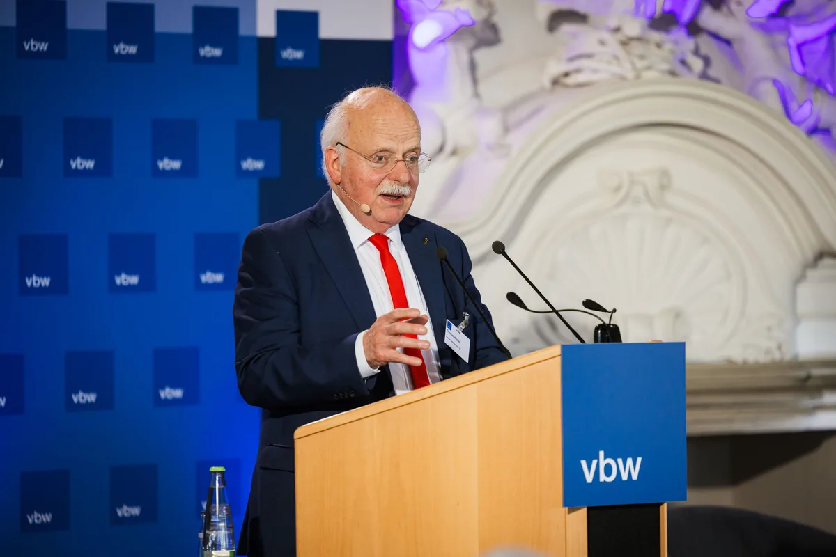 Älterer Redner mit Brille und roter Krawatte gestikuliert mit einer Hand am Pult, links die blaue Logowand