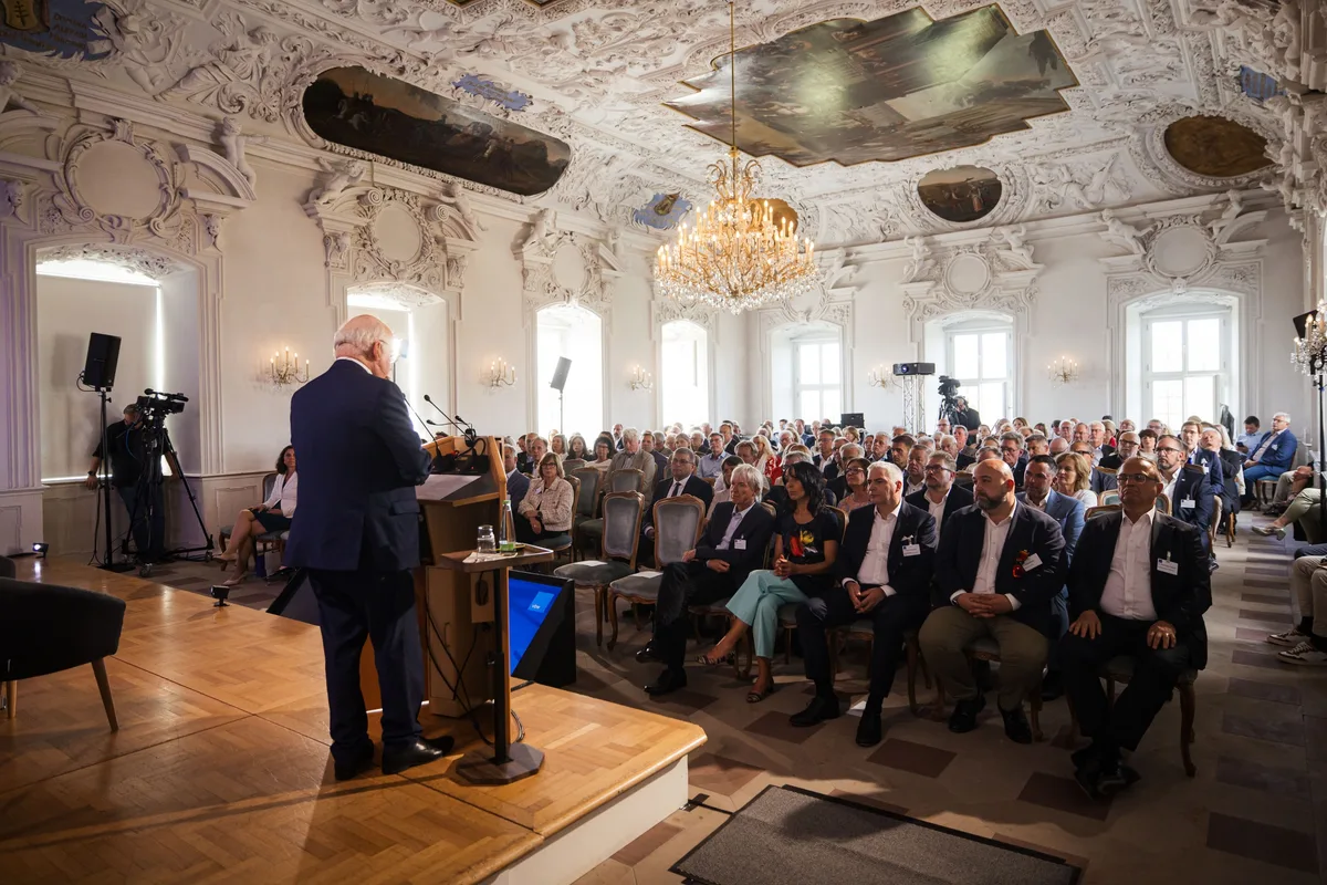 Redner von hinten am Pult auf dem Parkettpodium, vor ihm das Publikum unter Kronleuchter, links ein Kameramann am Stativ