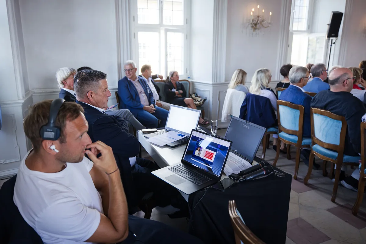 Mann im weißen T-Shirt mit Kopfhörer sitzt vor drei Laptops an einem Tisch, dahinter Zuhörer auf blau gepolsterten Stühlen