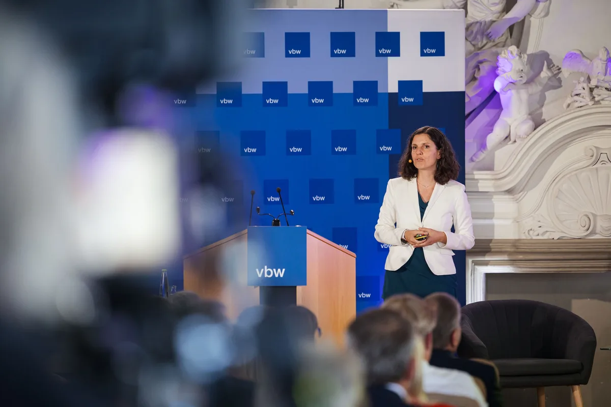 Vortragende in Weiß steht rechts vom Pult, im Vordergrund verschwommen eine Kamera und der Kopf des Kameramanns