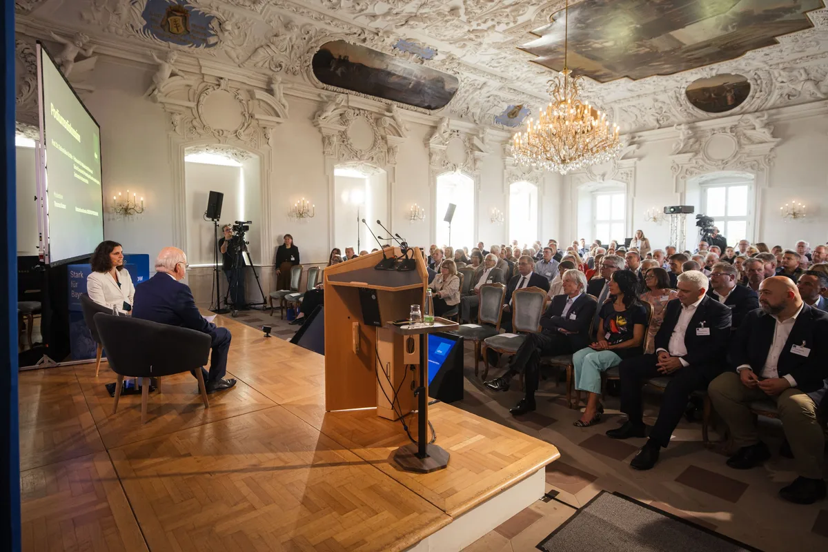 Auf dem Podium sitzen eine Frau und ein Mann in Sesseln für ein Gespräch, rechts das Publikum, links eine Leinwand