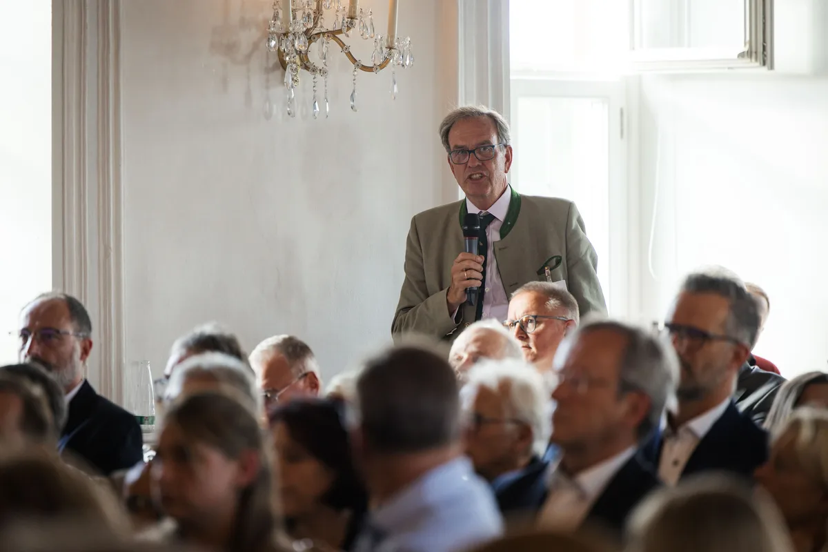Mann in grauem Trachtenjanker mit grünem Kragen steht im Publikum und spricht in ein Mikrofon, vor ihm Hinterköpfe