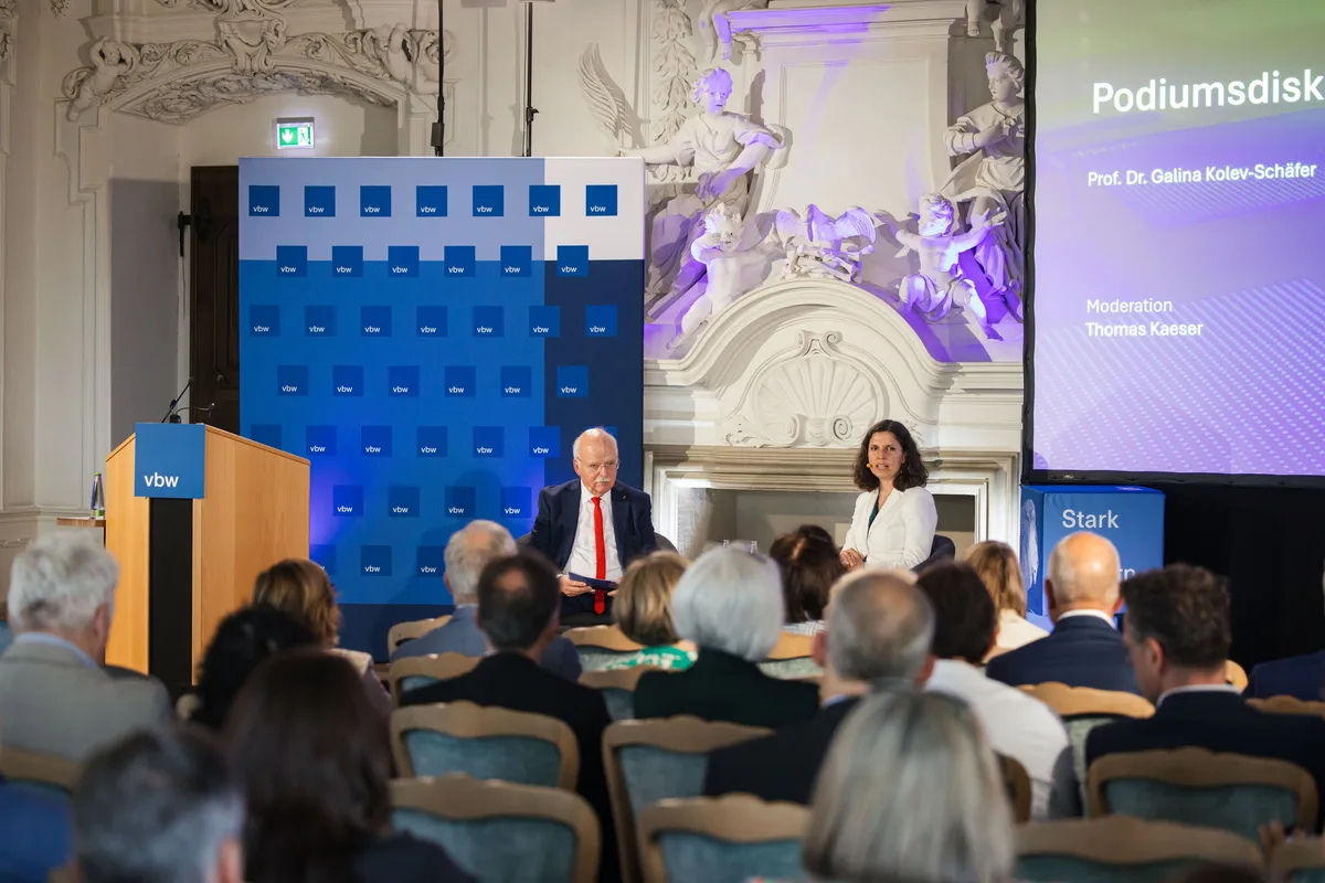 Gesprächsrunde auf dem Podium vor der blauen Logowand, links das Pult, rechts eine Leinwand, im Vordergrund Stuhlreihen