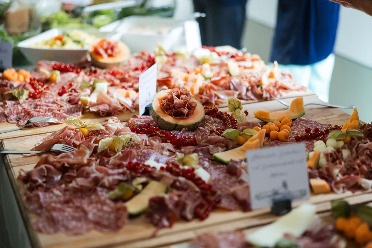 Holzbretter mit Aufschnitt, Schinken und Melonenspalten, dazwischen rote Johannisbeeren und Gabeln, dahinter Salatschüsseln