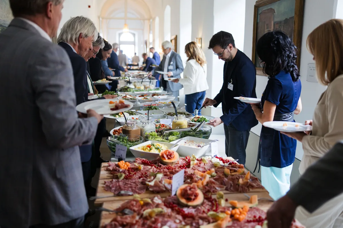 Gäste stehen mit Tellern am langen Buffet an, vorn Aufschnittplatten, dahinter Salate, hinten ein Flur mit hohen Fenstern