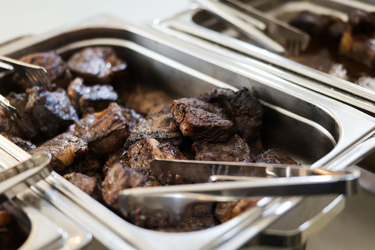 Gegrillte Fleischstücke dicht im Bild in einer Edelstahlwanne, eine Zange liegt quer darüber, daneben eine zweite Wanne