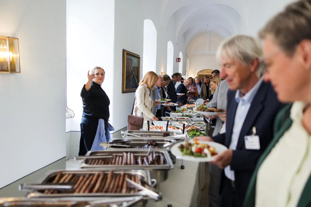 Gäste mit Tellern reihen sich am langen Buffet im Gewölbegang auf, vorn Wärmewannen mit Würstchen, links eine Servicekraft