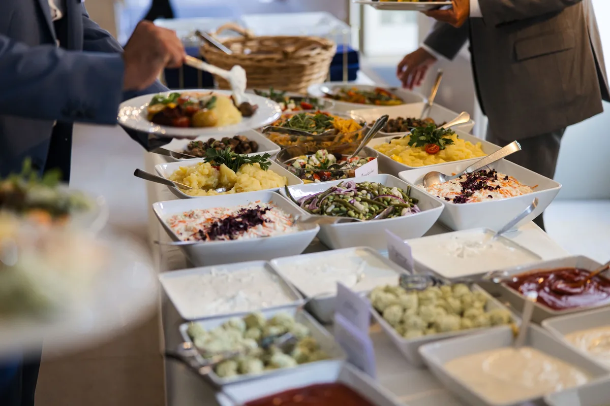 Weiße Schalen mit Kartoffel-, Bohnen- und Krautsalat sowie Dips auf dem Buffet, links und rechts füllen sich Gäste Teller