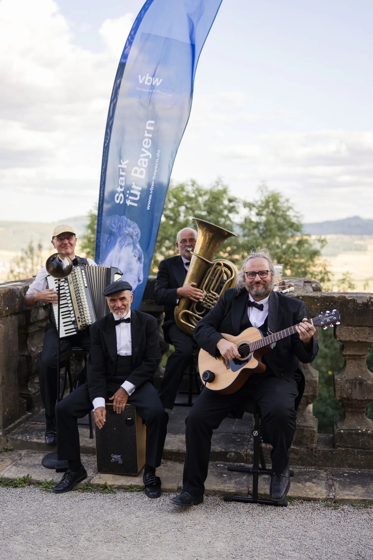 Hochformat: Vier Musiker in Schwarz mit Akkordeon, Tuba, Gitarre und Cajon vor einer Steinbalustrade, hinten ein Banner