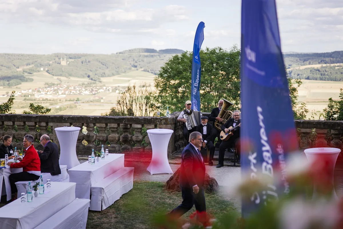 Auf der Terrasse spielt eine Band mit Tuba und Akkordeon, davor geht ein Mann im Anzug zwischen weißen Tischen durch