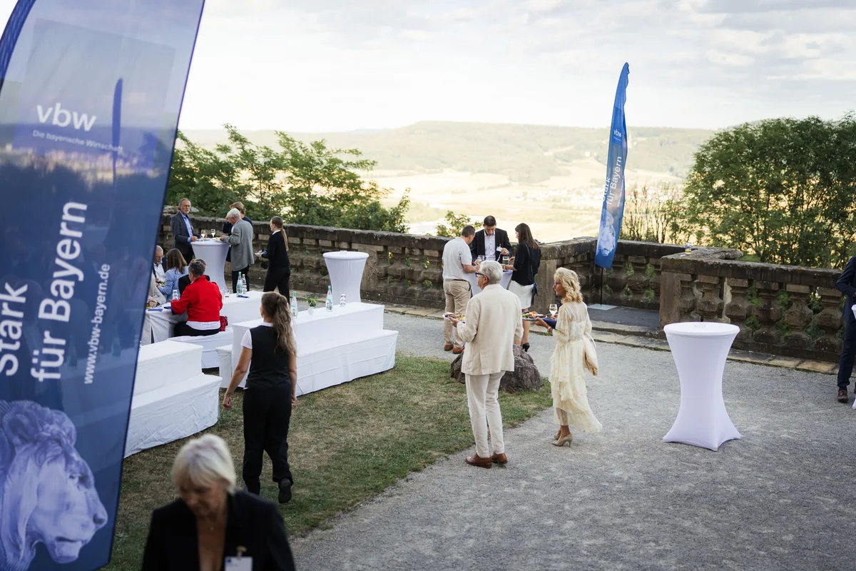 Blick von oben auf die Terrasse mit weißen Tischen, einzelne Gäste mit Tellern, links ein blaues Banner, hinten das Maintal