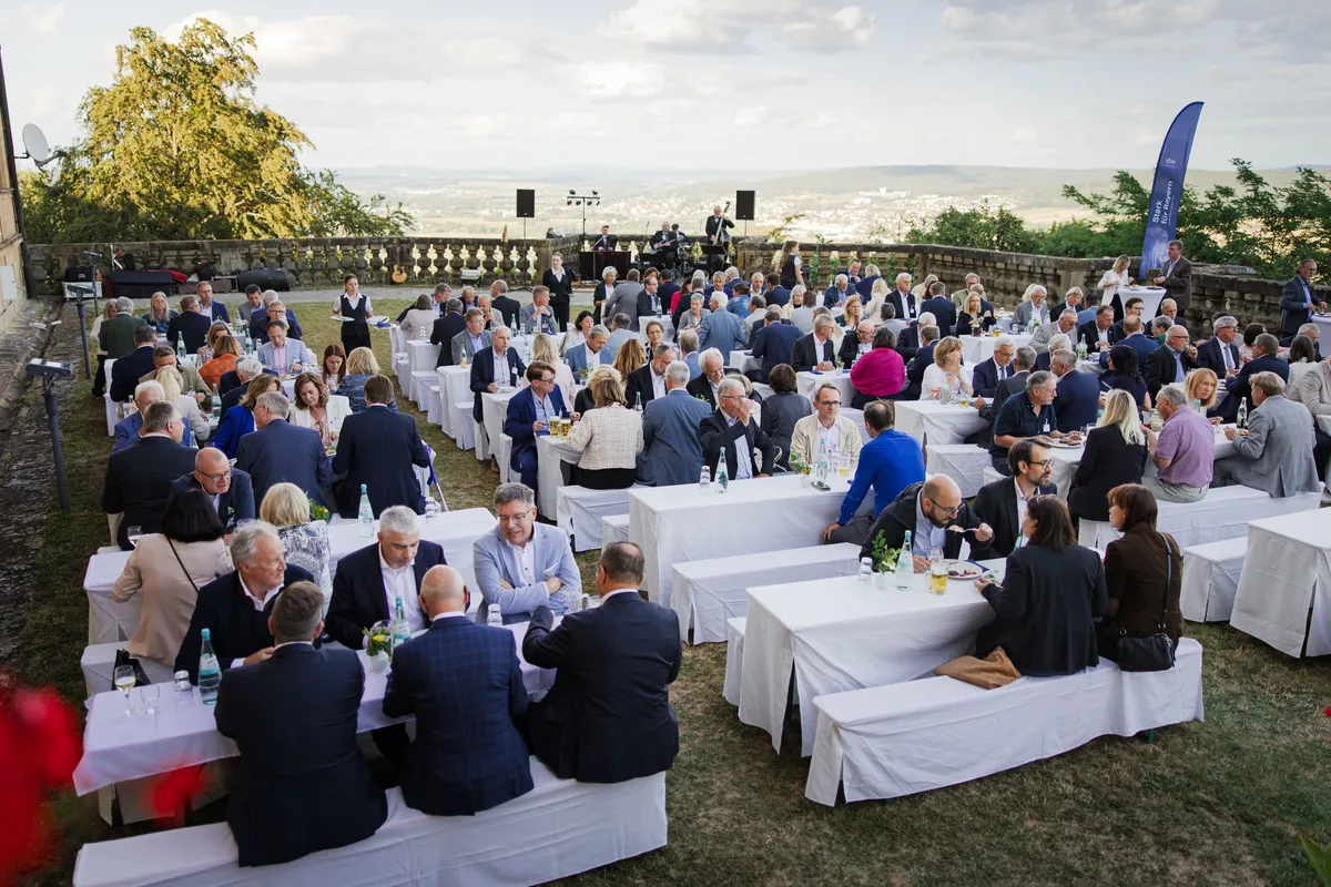 Terrasse am Abend voller Gäste an weißen Tischen mit Bänken, hinten die Band auf der Bühne, links Wiese und rechts ein Banner