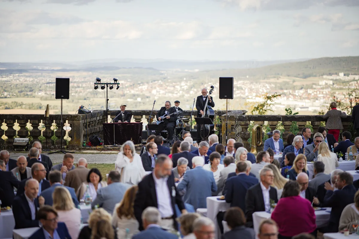 Die Band mit Kontrabass und Schlagzeug spielt auf der Terrasse, davor sitzen Gäste an Tischen, dahinter Hügel im Dunst