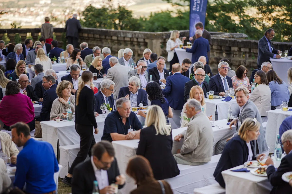 Gäste sitzen am Abend eng an weißen Tischen mit Gläsern und Tellern, hinten die Terrassenmauer und ein blaues Banner