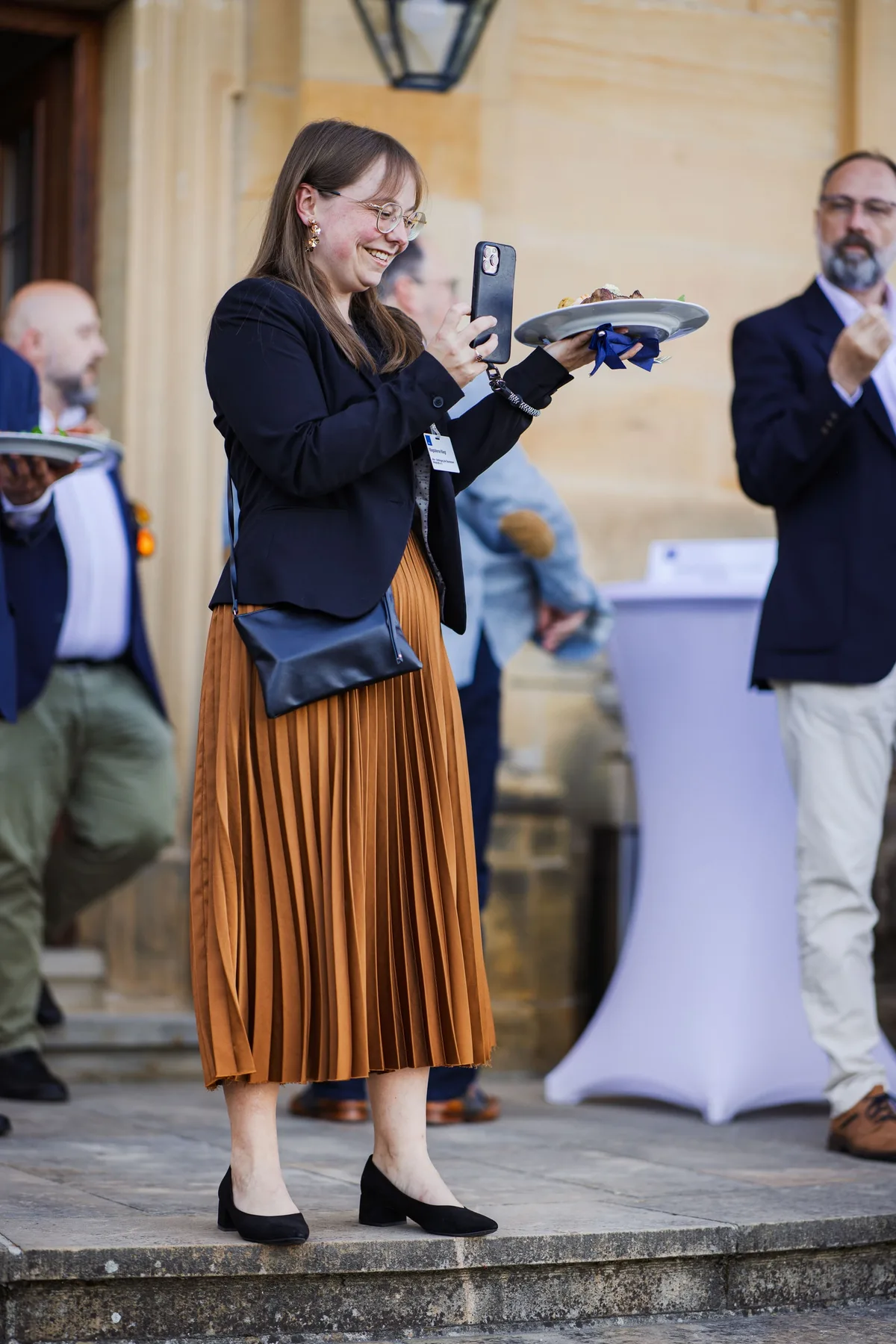 Hochformat: Frau in Plisseerock fotografiert mit dem Handy lächelnd ihren Teller, im Hintergrund Stehtisch und Sandsteinwand