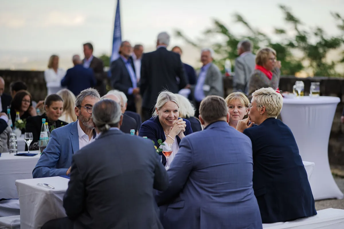 Gäste an Tischen auf der Terrasse in der Dämmerung, eine Frau im blauen Blazer lacht in die Runde, im Hintergrund Stehende