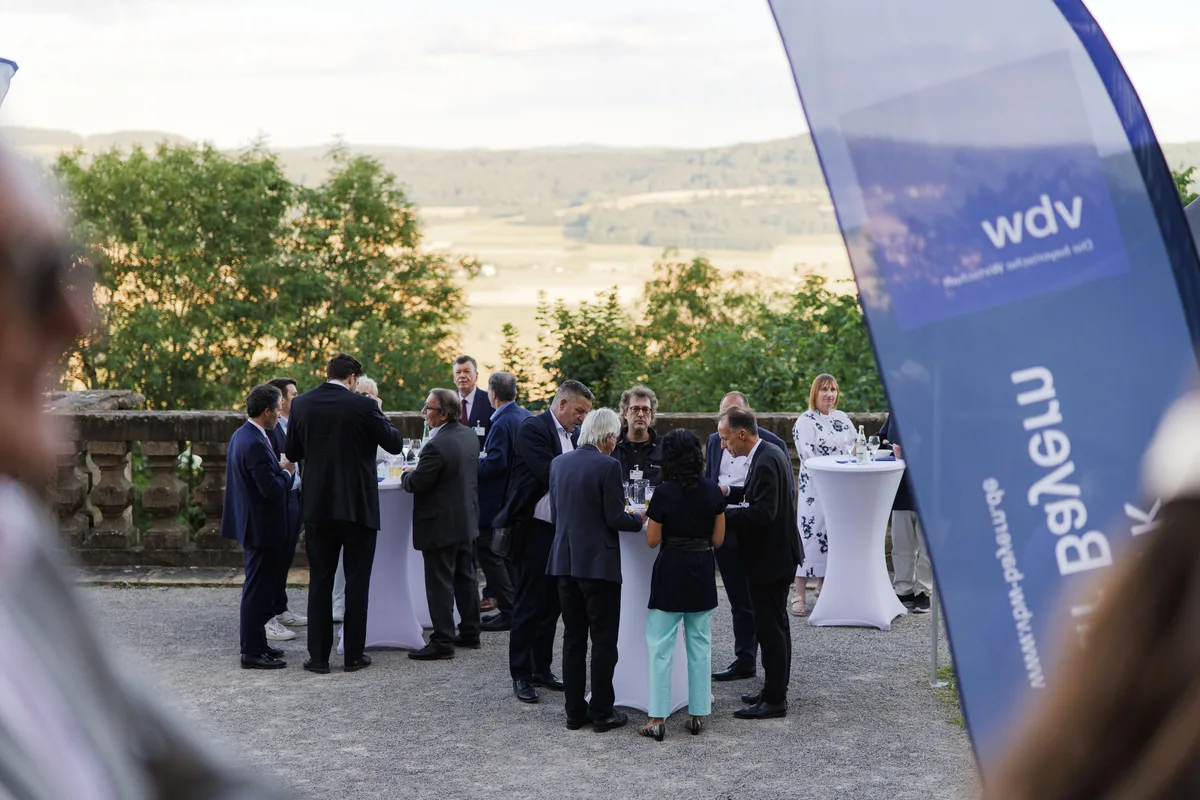 Kleine Gruppe von Männern und einer Frau steht an weißen Stehtischen auf dem Kies, rechts ragt ein blaues Banner ins Bild