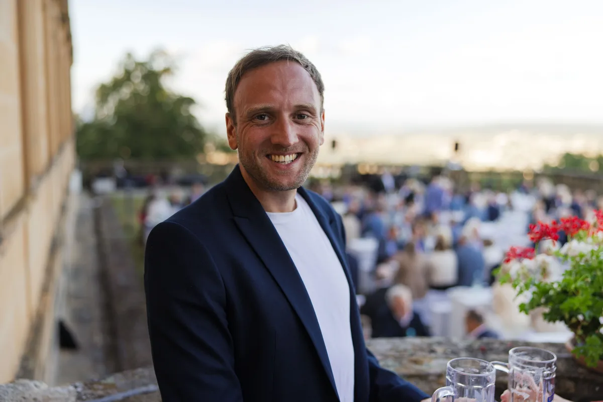 Mann im dunkelblauen Sakko mit weißem T-Shirt lacht in die Kamera, vor ihm zwei Gläser, dahinter die Gäste auf der Terrasse