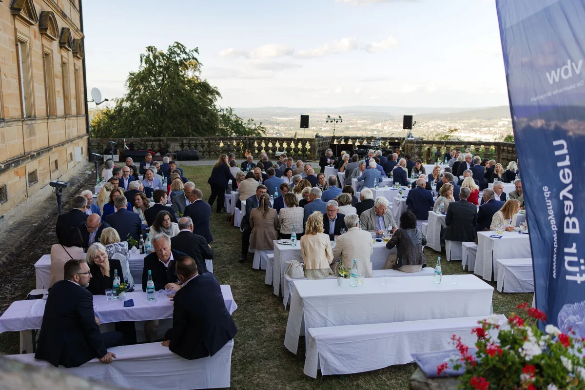 Terrasse am Abend mit langen weißen Tischen und Bänken auf dem Rasen, rechts ein Banner mit Geranien, links Sandsteinfassade