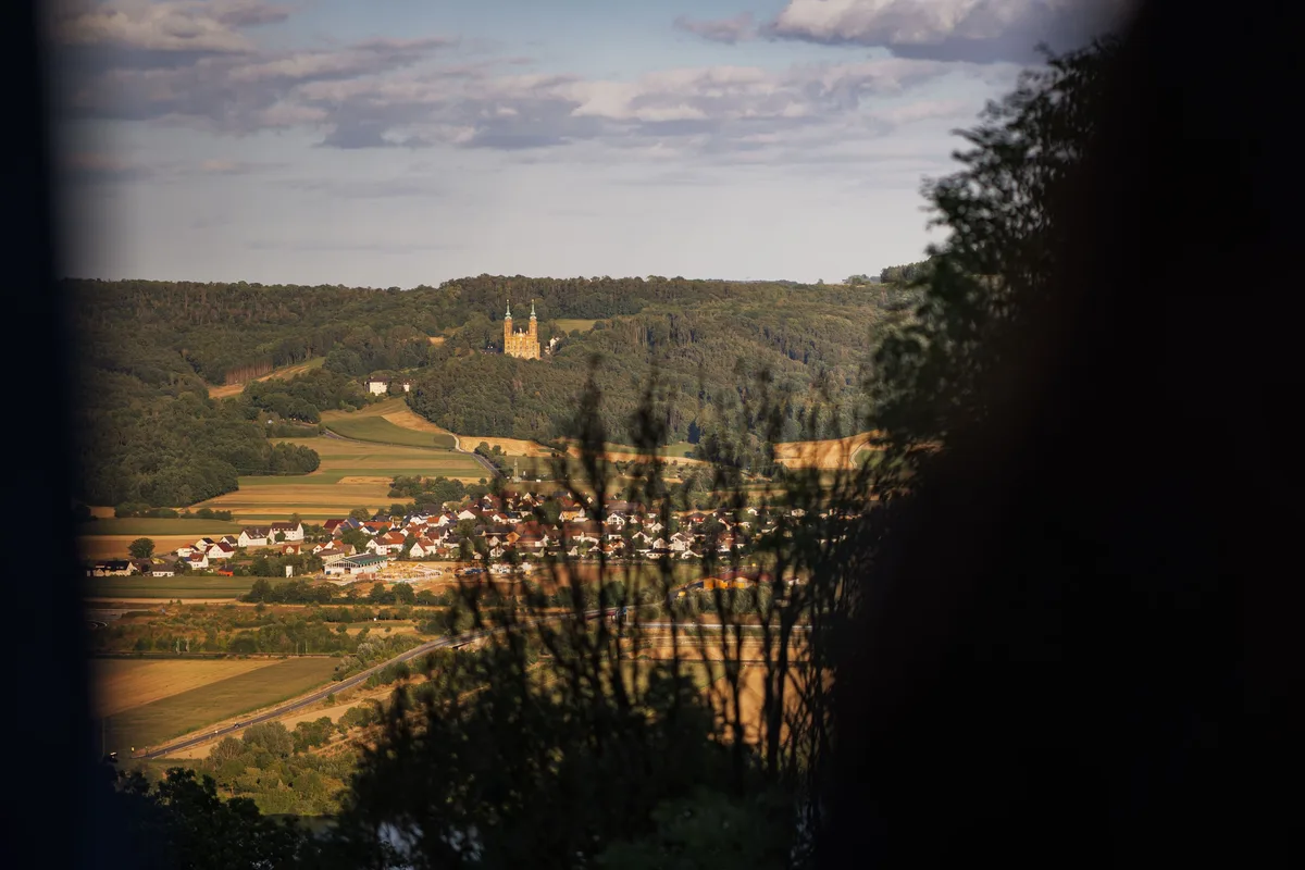 Blick durch dunkles Laub auf ein Dorf im Tal, dahinter ein bewaldeter Hügel mit einer Kirche mit zwei Türmen im Abendlicht