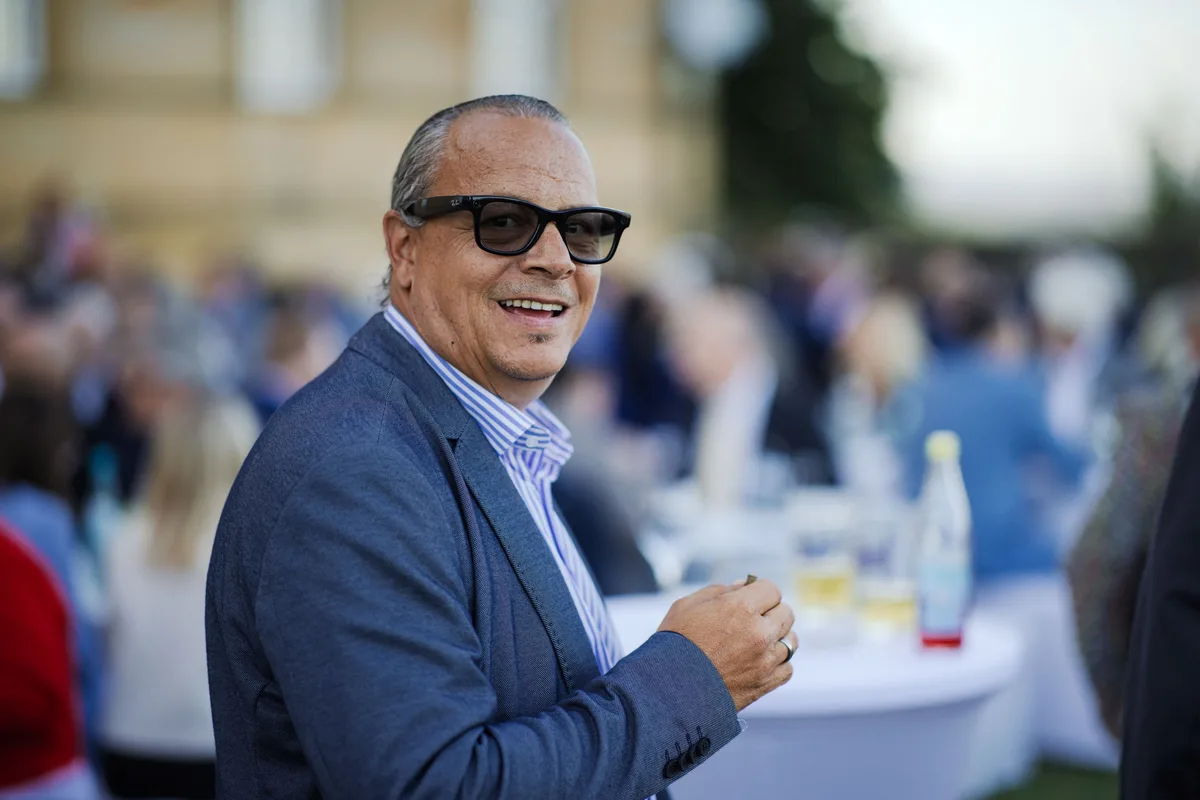 Mann mit dunkler Sonnenbrille und blauem Sakko lächelt zur Kamera, hinter ihm unscharf Stehtische und die Gäste auf der Wiese