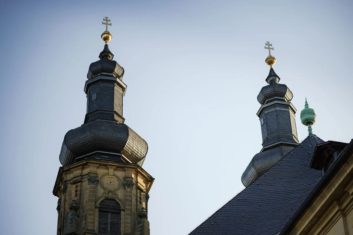 Zwei Kirchtürme mit Zwiebelhauben aus dunklem Schiefer und goldenen Doppelkreuzen vor blassblauem Himmel, rechts ein Dach