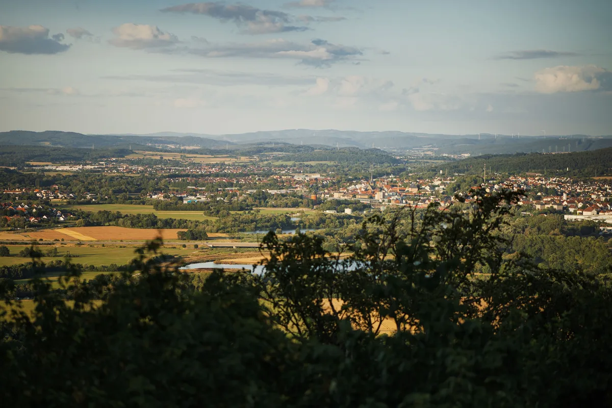 Blick aus der Höhe durch Laub auf eine Stadt im Tal mit Feldern, einem See und Hügelketten am Horizont
