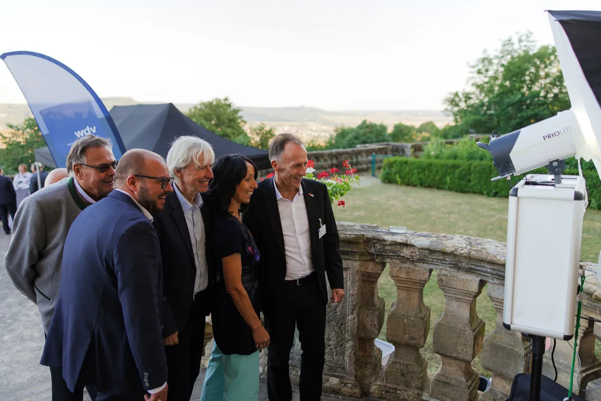 Vier Männer und eine Frau schauen lächelnd auf eine Fotobox mit Blitzlicht, die rechts an der Steinbalustrade steht