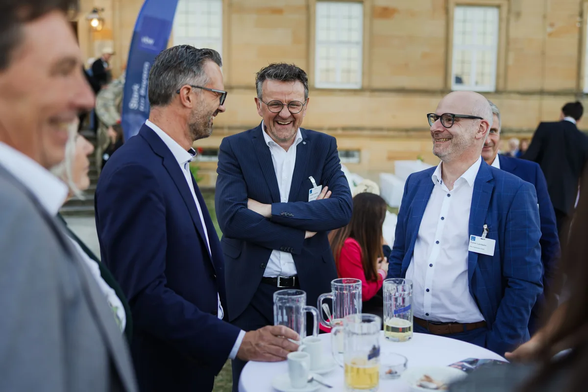 Drei Männer lachen im Gespräch an einem Stehtisch mit Bierkrügen und Kaffeetassen, hinter ihnen die Sandsteinfassade