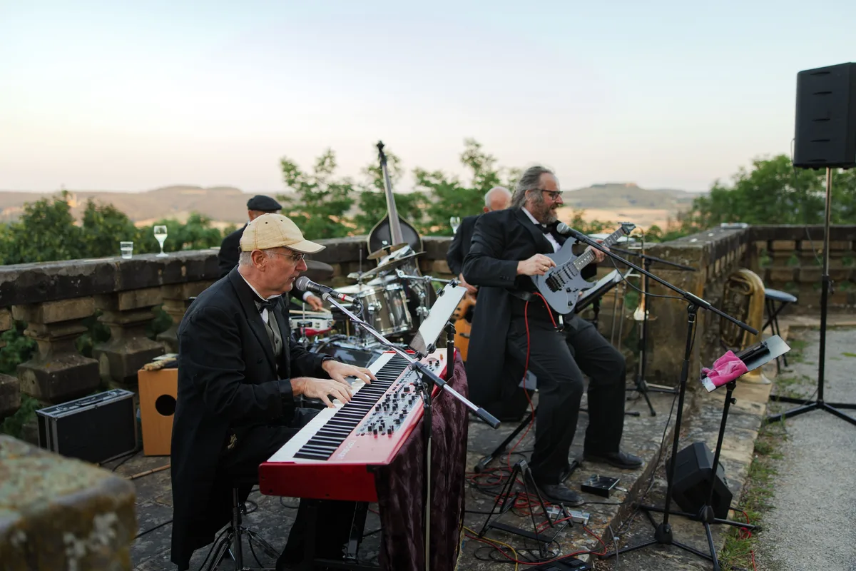 Keyboarder mit Schiebermütze und Fliege spielt neben einem Gitarristen im Frack, dahinter Schlagzeug und Kontrabass