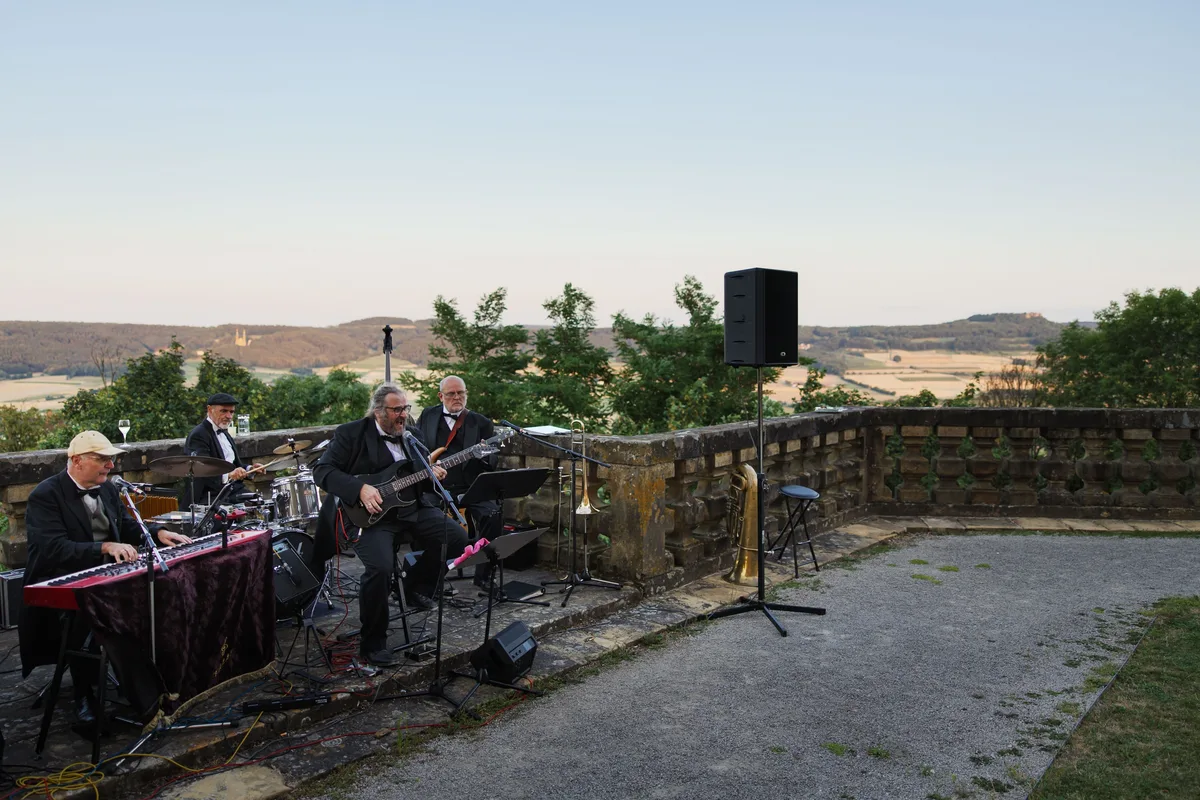 Fünfköpfige Band mit Keyboard, Gitarre und Schlagzeug spielt auf der Steinterrasse, rechts ein Lautsprecher, dahinter Felder
