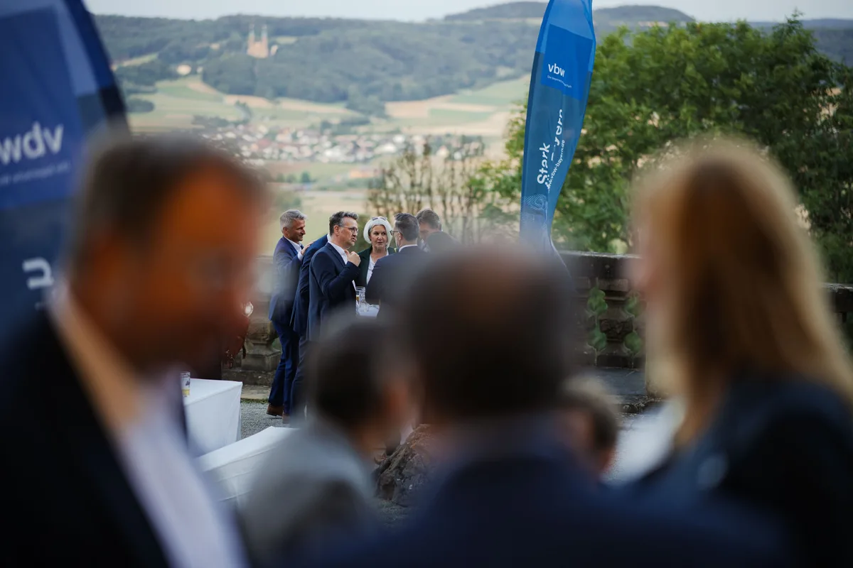 Unscharfe Köpfe im Vordergrund, dahinter eine Gesprächsgruppe an der Balustrade, über der ein Dorf mit Kirche im Tal liegt